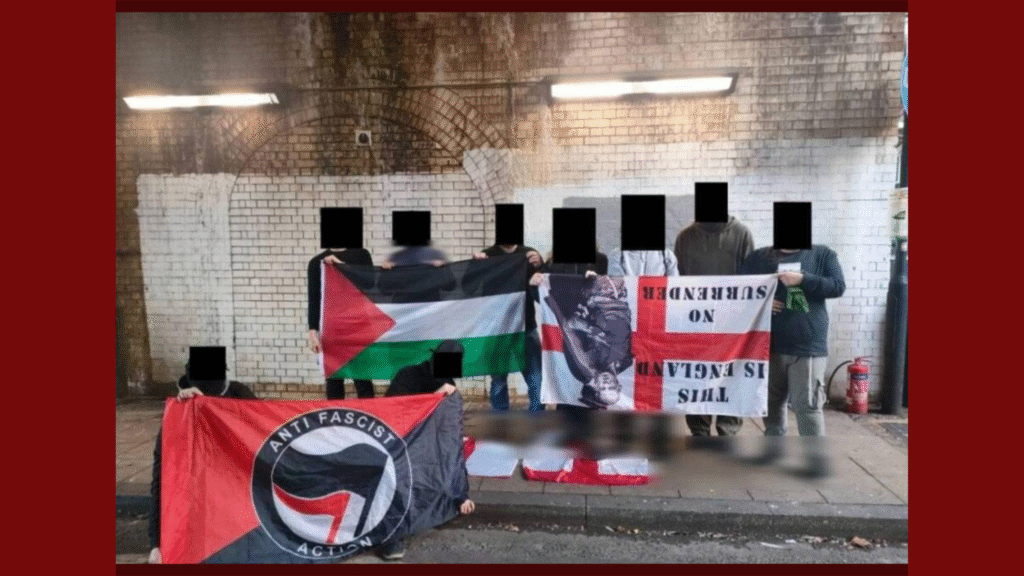 A group of people stand under a brick archway holding several flags. Faces and identifying features are blacked out. The flags include a Palestinian flag, an Anti-Fascist Action flag with a black and red circle logo, and a modified English flag with text reading “THIS IS ENGLAND NO SURRENDER.” The setting appears to be an urban street with a worn white-brick wall and overhead lights. 